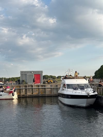 The image depicts a waterfront scene in Kastrup, Denmark, featuring a canal with several boats docked along a wooden pier. A modern white motor yacht with black trim is prominently positioned in the foreground, with several individuals visible on its deck. To the left, a smaller sailboat is moored, also with passengers onboard. Behind the pier, a grey building with a bright red door stands, and several bicycles and a yellow cargo bike are parked nearby. Trees line the background, and the sky is filled with a mix of clouds. The water appears calm, reflecting the surrounding structures and sky.