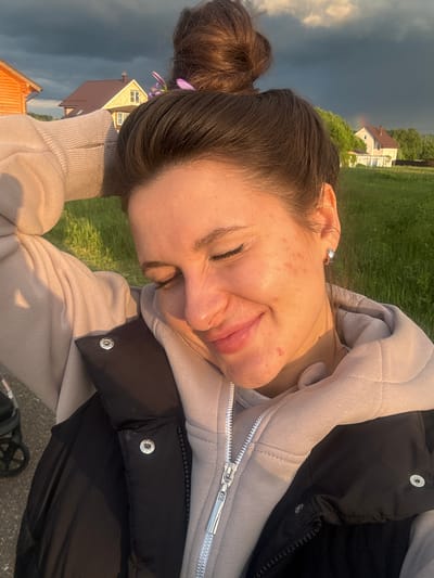 A sideways view of a woman lying on grass, with houses visible in the background. The woman has brown hair in a bun, adorned with purple flowers, and she wears a light-colored hoodie and a black vest. Her eyes are closed, and she appears to be smiling slightly. The location is inferred to be Vysokinichi, Russia based on other information not present in the visual image. The background shows a green field, houses with orange roofs, and a sky with ominous dark clouds. A rainbow is barely perceptible in the background. A stroller is present in the very lower corner of the image.