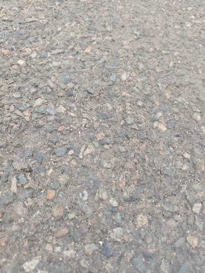 The image shows a close-up, overhead view of a rough, grey asphalt surface composed of small stones and gravel. There are no people or significant objects visible. The texture suggests it is an outdoor surface, possibly a road or pathway. The background is blurred, and there is a hint of dry vegetation in the upper left corner.