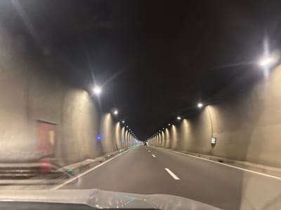 The image depicts the interior of a tunnel, likely a highway tunnel, with lighting fixtures mounted on the walls and ceiling. The tunnel has two lanes of traffic and is illuminated by bright lights. There are some blue and green lights visible along the sides of the tunnel. In the distance, a few vehicles are visible. The overall impression is that of a modern, well-maintained tunnel. The context provided mentions Surami, Georgia, suggesting this tunnel is located in or near that city.