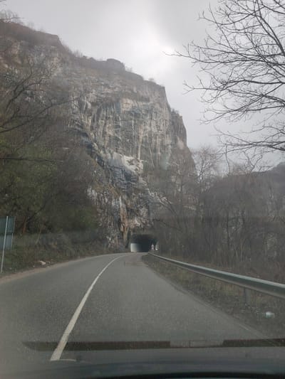 The image depicts a road leading towards a tunnel carved into a large rock face in Lyutibrod, Bulgaria. The sky is overcast. A two-lane road with a solid white line separating the lanes extends into the foreground. A metal guardrail is visible on the right side of the road. The rock face is a dominant feature on the left, with visible layers and textures. Sparse trees are visible along the sides of the road and on top of the rock face. A sign is visible near the tunnel entrance. The photograph appears to have been taken from inside a vehicle.