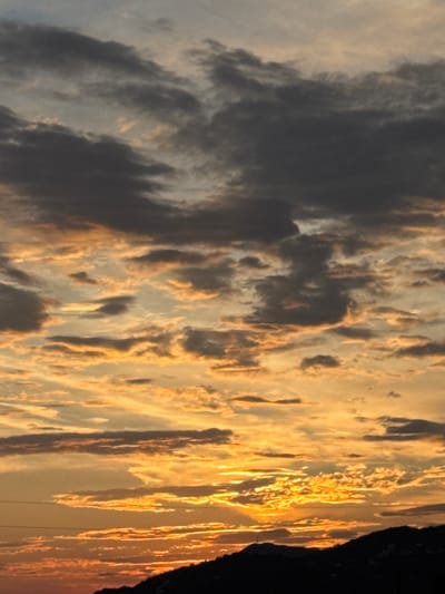 The image depicts a sunset over the city of Sustaš, Montenegro. The sky is filled with dramatic clouds, ranging in color from dark grey to vibrant orange and yellow, illuminated by the setting sun. The lower portion of the image shows a silhouette of a mountainous landscape, with hints of scattered lights suggesting the presence of buildings and possibly the outskirts of the city. The overall scene is one of natural beauty and the transition from day to night.