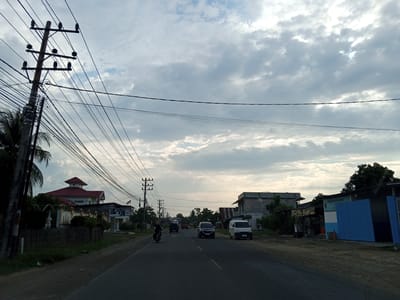 This image depicts a street scene in the city of Cotpuuk, Indonesia, under a cloudy sky. A paved road runs through the center of the frame, with a motorcycle and several cars traveling on it. On the left side of the road, a large utility pole with numerous wires is prominent, with a red-roofed building partially visible behind it. Further down the road, there are more buildings, including what appears to be a shop with a blue facade on the right. Trees and vegetation are present along the sides of the road, and the overall environment suggests a suburban or semi-rural area. The lighting indicates it is either early morning or late afternoon.
