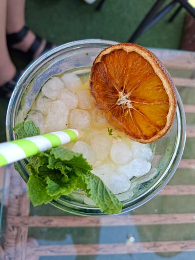 An overhead shot captures a tall glass filled with a light-colored beverage, ice cubes, and fresh mint leaves. A striped green and white straw is partially submerged in the drink, next to the mint. A dried orange slice garnishes the top of the beverage, positioned towards the right side of the glass. The glass sits on a transparent surface, likely a table, with a wooden slat pattern visible underneath. In the blurred background, a person's legs and feet wearing black sandals are visible, with a hint of green ground, possibly artificial turf, behind them. The scene is set outdoors, suggesting a relaxed, possibly cafe or bar environment in La Mata, Spain.