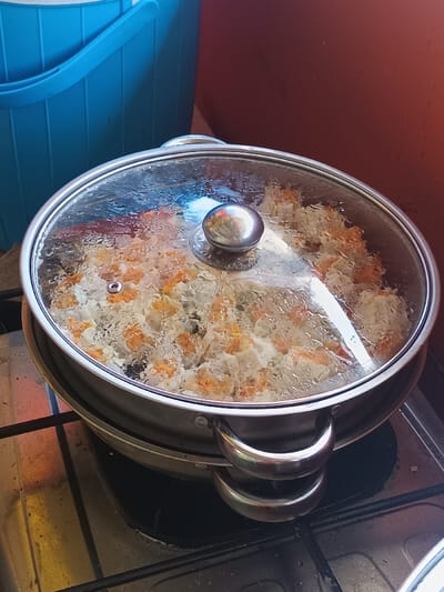 A steamer sits on a black stovetop, covered with a clear glass lid. Inside, there are numerous dumplings, identifiable by their white exteriors flecked with orange and dark specks. The steamer pot is made of stainless steel with looped handles. A blue bucket sits partially visible to the left of the steamer, and a reddish-brown wall is visible in the background. Condensation has formed on the inside of the glass lid.