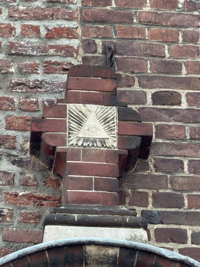 This is a close-up, eye-level, outdoor shot of a brick wall with a symbolic carving. The wall is constructed of red and darker red bricks with visible mortar. The carving, which is centrally located, is set into a lighter colored stone square and depicts an all-seeing eye within a triangle, surrounded by rays of light. This symbol is often associated with Freemasonry. The carving is mounted on a brick architectural element that juts out from the wall. The environment appears to be aged, with some weathering visible on the bricks and the carving. The lighting suggests it is daytime. The image captures a detail on the facade of the Munsterkerk in Roermond, Netherlands.