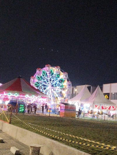 The image shows a carnival at night in Telukjambe, Indonesia, located at -6.307 latitude and 107.278 longitude. A brightly lit Ferris wheel dominates the center of the scene, with its colorful lights reflecting off the surrounding area.  A carousel, also illuminated, sits to the left of the Ferris wheel, with people standing near it.  There are various stalls and booths, all decorated with lights, suggesting a lively atmosphere. Several white tents are visible, likely providing shelter for vendors or other activities. People are scattered throughout the carnival, some standing, some walking, and some gathered around the booths and rides.  The area appears to be fenced off with yellow and black striped tape, suggesting it is a designated area for the carnival. The night sky is dark, punctuated by a few distant lights and stars.  A building with lit windows can be seen in the background.