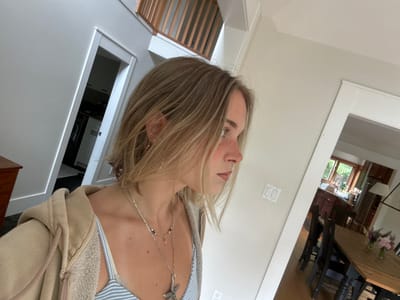 The image depicts a young woman in profile, positioned off-center to the left, looking towards the right side of the frame. She has light brown/blonde, shoulder-length hair and fair skin with visible freckles on her nose and cheek. She is wearing multiple layered silver necklaces, including one with a starfish pendant, and small stud and hoop earrings. Her attire consists of a light blue and white striped camisole or bralette, covered by a partially unzipped, light beige hooded sweatshirt with a textured inner lining.

The setting is the interior of a residential home, captured with a slightly wide-angle perspective. To the left of the woman, a light grey wall leads to a doorway with white trim, revealing a glimpse into a dark-floored room with white cabinetry and an appliance. Above this area, a wooden staircase with vertical balusters and a matching handrail ascends.

To the right of the woman, a neutral-colored wall features a white light switch plate. Further into the background, an open archway reveals a dining area with a light-colored wooden table and dark wooden chairs. On the table are two small clear vases containing pink and purple flowers. Beyond the dining area, a kitchen with dark wooden cabinets, a sink under a window, and a stainless steel refrigerator is visible. A lamp with a white conical shade hangs over the dining table. The floors in the visible areas are a mix of dark and lighter wood tones. The space is well-lit, suggesting natural light.