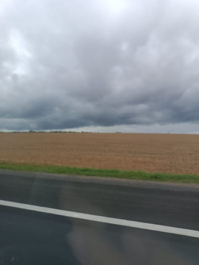An overcast sky looms above a flat, harvested agricultural field. In the foreground, a dark asphalt road with a white line is visible. The scene appears to be located in the rural landscape of Путрышкаўскі сельскі Савет, Belarus.