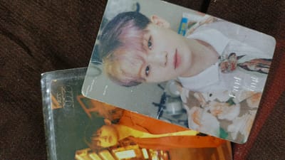 The image shows two K-pop photocards. One photocard, with a light-colored background and a person's face, is partially obscured by a second photocard. The second photocard is darker and shows the upper body of a person. Both photocards appear to be placed on a dark brown, textured surface, possibly fabric. There are no discernible elements indicating a specific location in Palohlada, Indonesia.