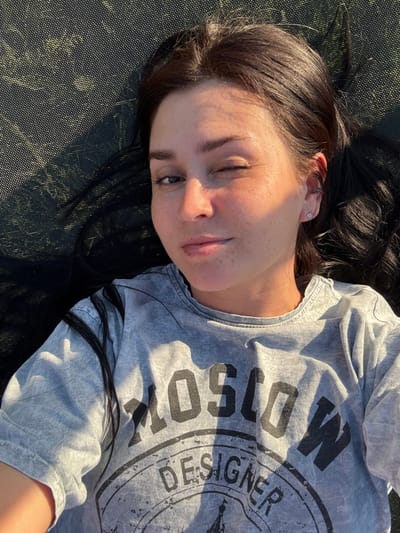 A young woman with dark hair is lying down outdoors, looking up towards the camera. She is wearing a grey t-shirt with "MOSCOW DESIGNER" printed on it in large, bold black letters. The woman's face is partially illuminated by the sun, casting shadows and highlighting her features. She has freckles across her nose and cheeks and is winking. Her hair is spread out around her head. The surface she is lying on appears to be a dark, textured material, possibly a trampoline or outdoor mat, with a slightly metallic sheen in places. The lighting suggests it is daytime, with the sun casting long shadows. The overall mood is relaxed and casual.