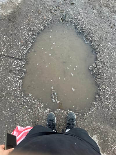 The media file shows a person standing next to a large pothole filled with murky water on an asphalt surface. The person is wearing black pants and black sneakers with white accents. A red and white striped bag is partially visible, held by the person. The person is holding a smartphone in their right hand, angled downwards. The pothole is irregular in shape, with jagged edges of broken asphalt and gravel. The water in the pothole is brown and has several small white bubbles on its surface. The surrounding ground is also asphalt, with loose gravel and some dry leaves. A thin stick or twig lies across the edge of the pothole. The lighting suggests it is daytime, and the overcast sky implied by the lighting and the presence of the puddle hints at recent or ongoing rain. The scene appears to be an outdoor urban or suburban environment, possibly a street or pathway with poor road maintenance. There is no visible text in the image.