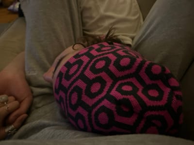 A single individual is depicted in a close-up, dimly lit indoor setting, appearing to be resting or asleep. The person's head, with dark curly hair visible, is turned to the left, revealing the profile of their nose and cheek. They are wearing a prominent fuchsia and black knit beanie with a repeating geometric hexagonal pattern. The individual wears light grey casual trousers, and a pale-colored shirt is visible underneath. A left hand, adorned with a silver-toned ring featuring a light-colored stone on the ring finger and another irregular metallic ring on the middle finger, rests on the grey fabric of the trousers in the foreground. The individual is reclined on what appears to be a light brown sofa or upholstered surface. The image does not contain any visual information to determine a specific location within Kingston, Canada.
