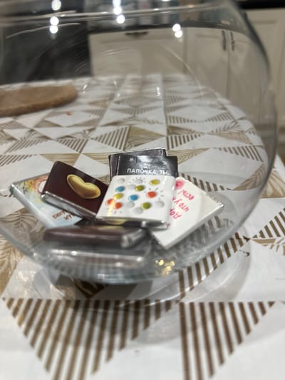 A close-up, eye-level shot captures a clear glass fishbowl filled with chocolates, sitting on a patterned tablecloth. The fishbowl's curved edge is visible in the foreground and to the right. Inside the bowl, several chocolates are stacked and arranged. One chocolate, prominently displayed, has a brown wrapper with a golden heart-shaped design on it. Adjacent to this is a white chocolate wrapper adorned with colorful dots in red, blue, yellow, and green. Another chocolate wrapper, partially obscured, features a floral design and Russian text. A dark chocolate wrapper is also visible. The tablecloth beneath the bowl has a geometric pattern of alternating white and brown stripes. In the blurry background, the interior of a room is suggested, with hints of a countertop and cabinetry. The lighting in the scene is bright, causing reflections on the glass surface. The image appears to have been taken indoors, in what could be a kitchen or dining area. The location is specified as Imeni Sverdlova, Russia.