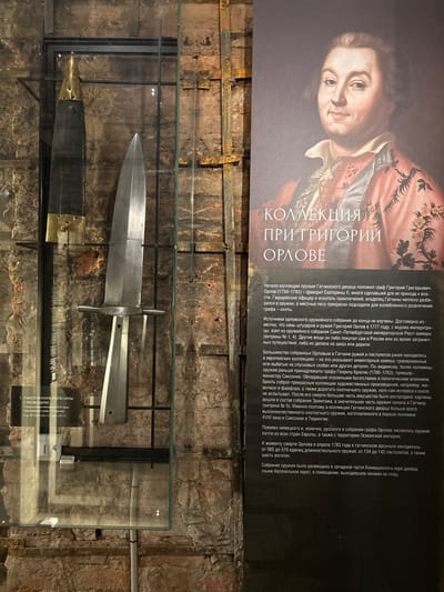 The image displays an exhibit in Gatchina, Russia, featuring historical weaponry. In the foreground, a glass display case contains two antique weapons: a dark, elongated object with a brass fitting at the top, and a large, metallic dagger with a crossguard. To the right of the display case is a poster with a portrait of a man in 18th-century attire and text in Russian, which translates to "Collection of Grigory Orlov." The text describes the origins and content of Grigory Orlov's weapon collection, noting its transfer to Gatchina in 1777. The background consists of a stone wall, suggesting an interior setting within a historical building. The overall scene appears to be a museum exhibit dedicated to a historical collection of arms and armor.