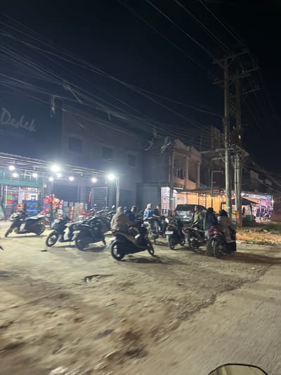 The image depicts a nighttime scene in what appears to be a commercial area or street in Bundar, Indonesia. The primary subjects are a line of parked motorcycles, with some people seated on or standing near them. In the background, there are buildings with some illuminated windows and a sign that reads "Dedek BSNL SHOP". The setting is characterized by the presence of numerous utility poles with tangled wires, suggesting an urban or semi-urban environment. The ground is a dirt or gravel surface, showing signs of wear. The lighting is artificial, coming from streetlights and building lights, indicating it is nighttime. There is no clear indication of weather, but the ground does not appear wet. The overall mood is one of everyday activity, likely people gathering or taking a break at a shop or establishment. The image's orientation is portrait, but the content is captured in landscape format, suggesting the photo was taken from a moving vehicle, possibly a motorcycle, looking towards the side.