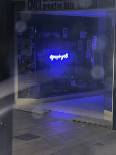 The image shows the exterior of a shop at night in Ibiza, Spain. The shop's name, "CMCI creativa ibiza," is visible above the entrance. Inside, a blue neon sign displays "@joaquinparli." The shop window reflects the blue light, and some items are visible within, including what appear to be framed pictures and musical instruments. The pavement outside is made of stone tiles, and a blue light from within the shop illuminates the ground. The scene is dark, with only the neon sign providing significant illumination.