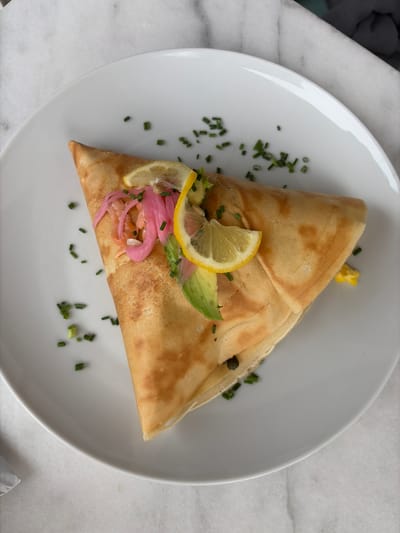 The image shows a folded crepe served on a white plate. The crepe is garnished with slices of lemon and avocado, pickled red onions, capers, and scattered chopped chives. The plate rests on a marble surface. The context suggests the photo was taken at a restaurant in College Station, United States of America.