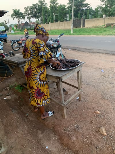 A woman in a yellow dress stands next to a wooden table holding a silver bowl filled with dark-colored meat. The woman is on the side of the road in Gbeleko, Nigeria. Several motorcycles are parked nearby. The ground is dirt and gravel.