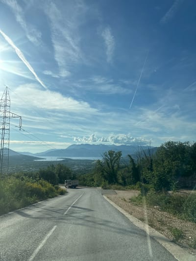 The image depicts a scenic view from a paved road in Pelinovo, Montenegro, on a bright, sunny day. The sky is predominantly blue with numerous white contrails, some distinct and linear, others more dispersed, indicative of aircraft activity. The sun, located in the upper left quadrant, casts a strong glare and lens flare.
In the foreground, a gray asphalt road with a white dashed center line curves gently downhill towards the center of the frame. The road's edges are lined with green foliage and some dry, lighter-colored vegetation. Shadows of unseen objects or trees are cast across the road surface, suggesting the sun is at an angle.
An electricity transmission tower, a tall metal lattice structure, is prominently positioned on the left side of the frame, extending vertically from the lower left towards the upper left. Multiple power lines run horizontally across the mid-ground from this pylon.
In the mid-ground, a light-colored truck, possibly a refuse or utility vehicle, is visible on the road, heading away from the viewer. The surrounding landscape consists of dense green trees and shrubs, covering the slopes and hills.
Further in the distance, a large body of water, likely a bay or a lake, is visible, reflecting the sky and appearing calm. Several small, indistinct boats or structures can be seen on the water. Beyond the water, a range of mountains with varied topography stretches across the horizon under a hazy sky, topped with a few distinct white clouds in the mid-right. No individuals are visible in the scene.