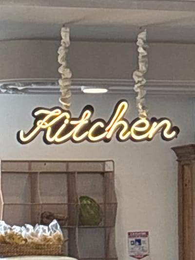 The image shows a neon sign that spells out "Kitchen" in cursive, illuminated with a warm yellow light. The sign is mounted vertically on a light gray wall. To the left of the sign, there are some white ruffled decorative elements hanging from what appears to be a shelf or rack. To the right of the sign, there is a multi-tiered shelf unit. On one of the shelves, a green, round object, possibly a watermelon or a large gourd, is visible. Further to the right, other decorative items with ruffles and textured fabric are present. In the upper right corner, a small sign with red and blue elements is affixed to the wall, though the text is too blurry to read. The lighting suggests an indoor setting, likely during the daytime or with artificial lighting that mimics daylight. There are no people visible in the image. The scene appears to be a display or decorative setup, possibly in a restaurant, cafe, or a home kitchen. The overall impression is one of a welcoming and perhaps rustic ambiance, with the neon sign serving as a prominent focal point. The location context provided is Rionegro, Colombia.