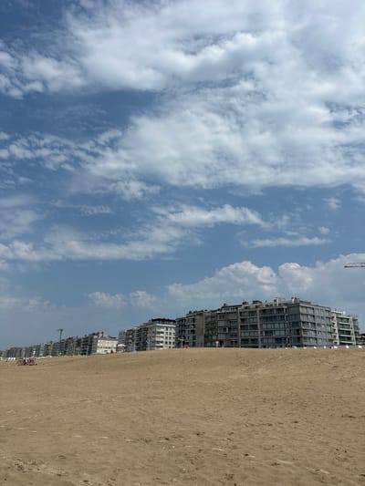 The image depicts a wide sandy beach in Koksijde, Belgium, with a row of modern, multi-story apartment buildings lining the coast in the background. The sky above is a clear blue, dotted with scattered white clouds. The beach is mostly empty, with a few people visible in the distance, some sitting on chairs or under umbrellas. The sand shows some footprints and natural undulations. The apartment buildings are characterized by large windows and balconies, suggesting a residential or holiday complex. The overall scene conveys a sense of a quiet day at the beach with urban development at its edge.