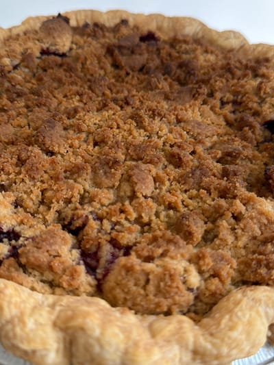 The image shows a close-up view of a baked pie with a golden-brown crumb topping and a light-colored pie crust. Darker purple filling is visible beneath the crumble. No people, actions, or interactions are depicted. The environment is not visible beyond the pie itself, and no location information is provided.