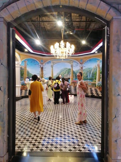 The image captures a view through an arched doorway into a room with an elaborate painted backdrop depicting a European landscape. The room's floor is covered in a black and white geometric tile pattern. A chandelier hangs from the ceiling. Several people are present, some facing away from the camera and seemingly engaged with a figure near the center of the room. One woman stands on the right side of the frame, wearing a floral dress and sandals, looking directly at the camera. Another person, dressed in a yellow garment, walks away from the camera. The scene is located within Hong Gai Ward, Vietnam.