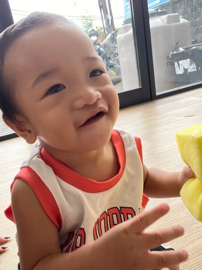 A baby of East Asian descent, approximately six to eighteen months old, is depicted in a medium close-up, smiling with an open mouth, facing slightly upwards and to the viewer's left. The baby has dark hair, dark eyes, and fair skin. They are wearing a white, sleeveless top with red trim around the neck and armholes; red lettering, partially obscured, is visible on the chest. The baby holds a bright yellow, soft, rectangular object in their right hand, while the left hand is open and extended. The baby is seated on a light brown wooden or laminate floor. In the lower-left corner of the frame, the tips of two fingers with dark painted nails are visible, suggesting the presence of another individual nearby.
The background reveals an outdoor scene through a large, dark-framed window, indicating the setting is indoors, looking out onto a street or yard in Uluwatu, Indonesia. Visible elements outside include several parked motorbikes, green foliage, utility poles with overhead wires, and a large, beige "Penguin" brand water storage tank. Adjacent to the water tank, a dark-colored bin is visible. The outdoor lighting appears diffused, suggesting an overcast day or shaded area.