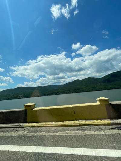 The image captures a scenic outdoor landscape in Drobeta-Turnu Severin, Romania, viewed through a window, likely from a moving vehicle. The scene is dominated by a clear blue sky with numerous scattered white cumulus clouds. The upper left portion of the sky exhibits a prominent diagonal light flare or reflection, along with indistinct lighter shapes, suggesting reflections from the interior of a vehicle. Two distinct, bright, small rectangular reflections are visible in the upper right sky area.
Below the sky, a vast body of dark blue-grey water extends horizontally across the mid-ground. Beyond the water, a series of lush green, densely forested mountains or hills rise, forming a continuous undulating horizon line across the distant background.
In the foreground, a concrete barrier is visible. The primary section of the barrier is painted a bright yellow, with a small square, yellow-capped post-like structure positioned towards its left. To the immediate left of this post, the barrier appears to be unpainted grey concrete. Below the main yellow barrier, a narrower yellow curb or ledge runs parallel. The very bottom of the image shows a section of dark asphalt road, featuring a thick, solid white line running horizontally, indicative of a lane marker. No people or animals are visible in the scene, and no specific actions are occurring. The environment suggests a sunny day with good visibility, highlighting the natural beauty of the region alongside man-made infrastructure.
