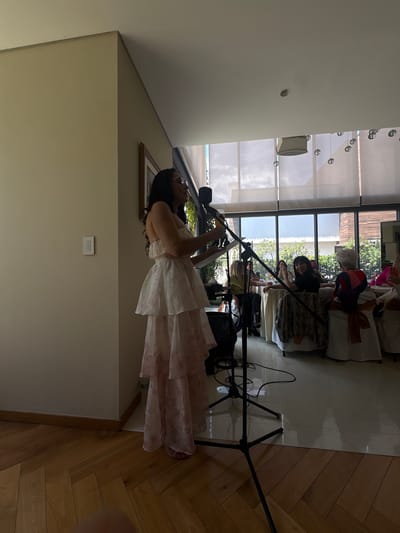 The image shows a woman in a light-colored, ruffled dress standing and looking at a tablet or book in her hands. She is positioned in the foreground of the image, with a microphone stand and audio equipment in front of her. In the background, through a large window, several other people are seated at tables, suggesting an event or gathering. Some of these individuals appear to be women wearing festive clothing. The setting appears to be indoors, possibly a venue or restaurant, with natural light coming from the windows. The time of day seems to be daytime, given the lighting. The woman in the foreground has dark hair and glasses. The overall atmosphere suggests a formal or semi-formal occasion, perhaps a performance, presentation, or celebration. There is no visible text in the image.