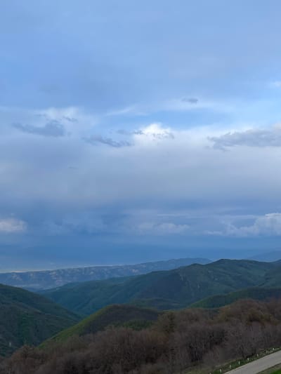 The image depicts a mountainous landscape under a cloudy sky in Zemo Nichbisi, Georgia. A range of green mountains occupies the right side of the frame, with a glimpse of a road on the far right edge. The sky, predominantly filled with light blue and grey clouds, takes up the majority of the image on the left side. The scene is shot from an elevated perspective, capturing the scale of the mountains and the vastness of the sky.