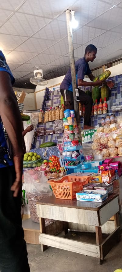 In Aviation Quarters, Nigeria, a young man is perched on a makeshift ladder made of wood, reaching for a large green papaya hanging from a shelf. He wears a dark purple t-shirt and dark pants. The shelves behind him are laden with various goods: bottles of what appear to be oil or sauce, bags of snacks, and assorted packaged food items, including Bounty chocolate bars. To the left, another person, only their arm and shoulder visible, is wearing a blue t-shirt. A white oscillating fan is mounted on the ceiling above. The overall setting is a small shop or market stall, with shelves constructed from wood and a tiled blue and white backsplash. The floor is concrete.