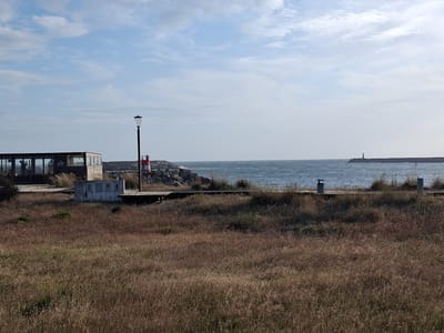 The image depicts a coastal scene in Figueira da Foz, Portugal. In the foreground is a field of dry, brown grass. Behind the grass, a wooden boardwalk runs along the coast. Several structures are visible along the boardwalk including a small, enclosed building with windows, a lamppost, and a concession stand. Beyond the boardwalk and structures, there is a rocky breakwater with a red and white marker. In the background, the ocean stretches to the horizon, with a distant structure visible in the distance. The sky is partially cloudy with blue patches.