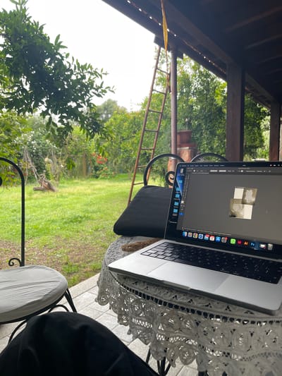 The image shows a laptop computer on a white lace-covered table. The screen displays a design software interface with a tile pattern. The laptop rests on a black cushion placed on a chair. The setting appears to be an outdoor covered patio or porch, with a garden visible in the background, including a ladder. The vegetation and architecture suggest a rural location in Italy, specifically Muristenis/Monastir. Another chair is visible with a white cushion. A person is partially visible in the frame, wearing black clothing, on the chair beside the laptop.