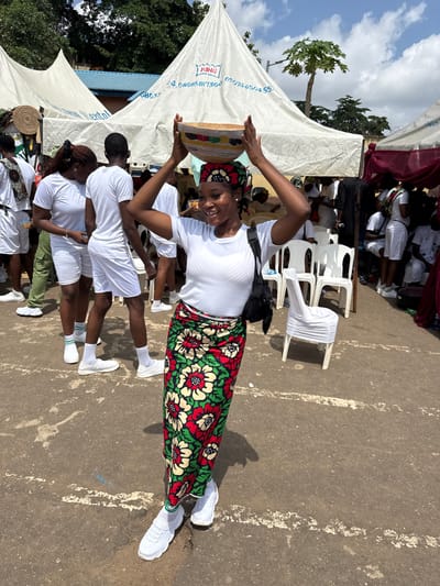 In Alagba, Nigeria, a young woman in a white t-shirt and a colorful floral skirt balances a decorated bowl on her head. She is smiling at the camera and her right hand is raised, supporting the bowl. She wears a headscarf that matches her skirt. White tents with blue lettering are set up behind her, one displaying the word "KING." Several people in white t-shirts and shorts are visible in the background, some walking and others seated on white chairs. The ground is paved with asphalt and has some white markings. Trees and a blue roof are visible behind the tents. The sky is partly cloudy.