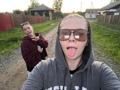 Two young women are captured in a selfie in what appears to be a rural village setting. The woman in the foreground is wearing a grey hooded sweatshirt and large, square sunglasses with a tortoiseshell pattern. She is sticking her tongue out, and her head is tilted slightly. The woman in the background, to the left, is wearing a dark maroon hooded sweatshirt and has sunglasses perched on her head. She is also sticking her tongue out and making a "rock on" hand gesture. Both women have light skin and blonde hair.

The environment is a dirt road lined with grass and gravel on both sides. Behind the women, there are wooden fences, and in the distance, several rustic wooden houses with dark roofs are visible. The sky is overcast, suggesting it might be dusk or dawn. The overall impression is of a casual, playful moment shared between friends in a country setting. The location is identified as Borodulino, Russia.