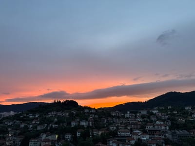 The image depicts a panoramic view of the city of Momchilovtsi, Bulgaria, during twilight. The sky is filled with a gradient of colors, transitioning from a soft lavender and gray at the top to vibrant oranges and pinks near the horizon. A band of darker clouds stretches across the sky, partially obscuring the setting sun.

Below the sky, the scene is dominated by a densely populated hillside. Numerous houses with red-tiled roofs are nestled closely together, their white and light-colored facades contrasting with the dark green of the surrounding trees and mountains. The architecture appears traditional, with many buildings featuring balconies and multi-story structures. The hillside is steep, indicating the topography of the region.

The foreground shows the upper portions of buildings, suggesting the vantage point is from a higher elevation, looking down into the town. Some lights are visible in the windows of the houses, indicating that it is evening and some residents are home. The overall impression is one of a tranquil, mountainous village bathed in the warm, fading light of sunset. The vegetation is lush and green, typical of a mountainous area. The image captures a serene and picturesque moment in this Bulgarian town.