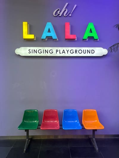 The image depicts a wall with colorful, three-dimensional letters spelling out "L A LA" and "oh!". To the right of the letters is a vertical, elongated white sign with black text that reads "SINGING PLAYGROUND". Mounted on the wall to the right of the sign are four brightly colored plastic seats stacked vertically: orange, blue, red, and green. These seats appear to be simple, molded plastic chairs, each attached to a black metal bracket that secures them to the wall.

The background wall is painted in a gradient of purple and grey. There are subtle shadows cast by the letters and the seats, indicating a light source from above and to the left. The overall impression is a playful and inviting space, likely designed for entertainment or a creative activity. The scene appears to be indoors, possibly in a public space like a community center, entertainment venue, or a creative studio. There are no people visible in the image. The lighting suggests daytime or well-lit indoor conditions. The location context of Incheon, South Korea, does not add any specific visual cues to the image itself.
