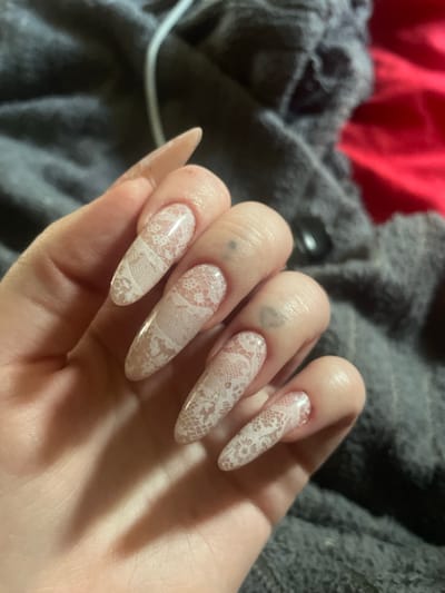 The image shows a close-up of a person's hand with long, almond-shaped nails decorated with a white lace pattern. The nails are polished and appear to be gel or acrylic. The hand is resting on a dark gray, fuzzy fabric, possibly a blanket or clothing. In the background, a portion of a bright red fabric is visible. The lighting suggests an indoor setting. There are no visible landmarks or details that would indicate the location in the city of Линёвка, Russia. The image focuses solely on the hand and nails.