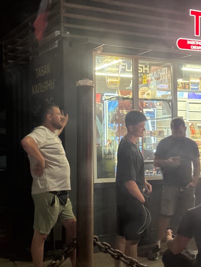 The image depicts an outdoor scene at night, likely in Kagal'nitskaya, Russia, with a small storefront or kiosk in the background. The kiosk has large glass windows displaying various products, including what appears to be tobacco and electronic cigarette items, with signage in Russian. Three men are visible in the foreground and midground.
On the left, a man in a light-colored t-shirt and light green shorts is standing and gesturing with his right hand. He has a pouch or bag tucked into the waistband of his shorts. In the center, a younger man in a dark t-shirt and dark shorts stands facing towards the kiosk, holding a white cord or wire. To the right, a third man in a dark t-shirt and light-colored shorts is holding a smartphone in his right hand, looking down at it. Another individual's arm and torso are partially visible on the far right, seemingly holding a cigarette. A metal chain is in the lower foreground, and a metal pole is to the left of the man in the light t-shirt. The lighting is artificial and casts shadows, indicating it is nighttime.