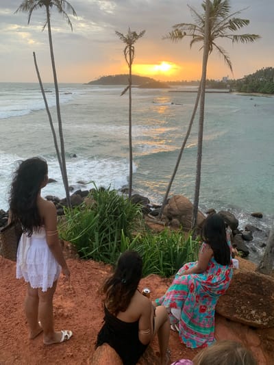 The image depicts a sunset scene at Thalaramba, Sri Lanka, with three women observing the ocean from a cliff. In the foreground, a woman in a white dress stands facing the sea, while another woman in a floral dress is seated on a rock. A third woman, wearing a black top, is also seated. Palm trees are scattered across the landscape, and the ocean features gentle waves. The sky is partly cloudy, with the sun setting on the horizon, casting a warm glow on the water. The environment is coastal, with rocky outcrops and lush vegetation.