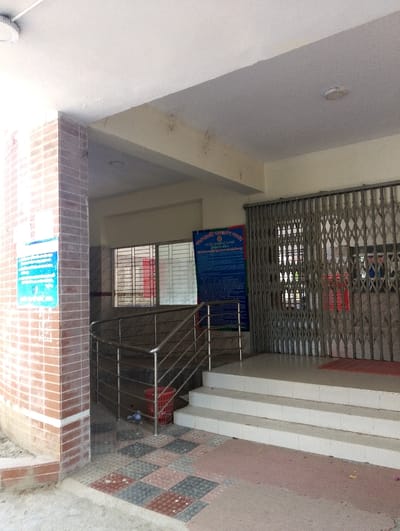 The image shows the entrance to a building in Bhola, Bangladesh (latitude: 22.688, longitude: 90.622). There are four white steps leading up to the entrance. To the left is a red brick wall and a stainless steel handrail leading down to a ramp. The entrance is protected by a metal gate that is shut. There is a blue sign on the wall to the left with white text that is mostly obscured. To the right of the entrance is a poster with white text on a blue background. The poster is secured to the wall with clear tape. There are three light fixtures visible in the ceiling. The floor is tiled, and there is some dirt and debris on the ground in front of the ramp. The image was taken during the day, and there are no people in the image. The building looks to be in an urban area.