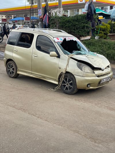 The image shows a wrecked, gold minivan, with the windshield shattered and the front hood severely damaged, parked on a paved road in Githurai, Kenya, at the coordinates -1.188, 36.933. The vehicle is facing the camera. The front passenger-side window is broken and there are several items of rope hanging off the vehicle, likely from the wreckage. The road appears to be a local street with a small amount of traffic in the background. The sky is grey and overcast. The road is unpaved, likely dirt or gravel.