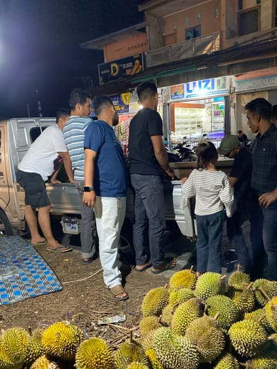 This media file captures a nocturnal scene in Lambheu, Indonesia, where a group of people are gathered around a stall or vehicle laden with durians. The primary subjects are the individuals, whose faces are mostly obscured or turned away, engaged in apparent interaction with the durian seller or examining the fruit. The setting appears to be a roadside stall or a temporary setup, indicated by the dimly lit background featuring storefronts with signage and displays of goods.
The prominent objects in the scene are the numerous durians, piled high and clearly visible due to their distinctive spiky exteriors. A vehicle, likely a truck or van, is partially visible, suggesting a mobile stall or a mode of transport for the fruit. Several people are standing and observing the durians. One person in the foreground is wearing a blue t-shirt and jeans, while another in a white t-shirt and black shorts is also visible.
The time of day is clearly night, given the darkness of the surroundings and the artificial lighting. The weather is not discernible from the image. Among the visible text, "DIARIM", "15.000", "FS LUZIA", "PARFUME", "Lampu Merah Keutapang", "TOKO MAS CAHAYA", and "JL. WEDANA/SIMPANG KEUTAPA" are present on various signs, suggesting the commercial nature of the location. The overall mood appears casual and transactional, typical of a nighttime fruit market.