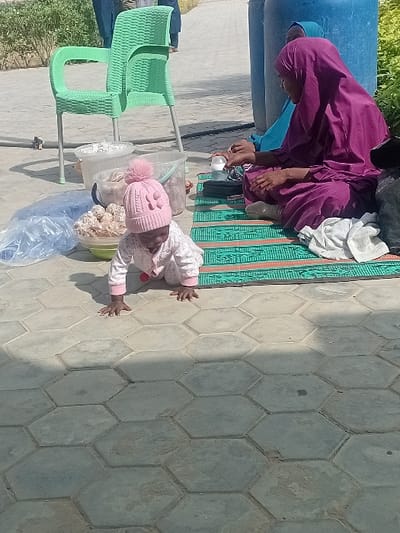 The scene is an outdoor setting in the city of Kwazailewa, Nigeria, under direct sunlight, indicated by strong shadows on the hexagonal grey concrete pavers. In the foreground, an infant is crawling on the pavement, dressed in a light-colored long-sleeved outfit and a pink knitted hat with a pom-pom. To the right, a woman in a dark purple hijab and matching long dress sits cross-legged on a green and orange patterned mat, holding a clear baby bottle with a white cap. A second woman, wearing a light blue hijab, is partially visible behind the first, also seated on the mat. Adjacent to the mat and the infant, an empty light green plastic chair with a woven pattern stands on the pavement. Near the chair are two translucent white plastic buckets, one containing unidentified light-colored contents, and a light-colored bowl filled with numerous small, spherical, light-colored items, possibly food products. A transparent blue plastic bag rests on the pavement near the bowl. Behind the seated women, two large blue plastic barrels are visible, partially obscured. In the distant background, two indistinct figures walk away from the viewer on what appears to be an unpaved area with some sparse green vegetation.