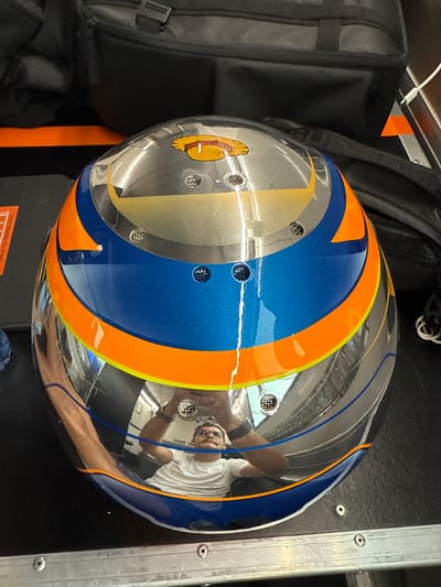 The image shows a blue, orange, and silver helmet sitting on a gray, metal surface. The helmet has a reflection of a man with glasses and a white shirt. He is holding something in his hand and it is unclear what he is doing. The helmet is sitting on top of a black case that is partially visible in the top of the frame. The surface the helmet is sitting on has multiple screws. The scene appears to be inside a garage or workshop. The location based on coordinates (latitude: 41.57, longitude: 2.259) is Montmeló, Spain.