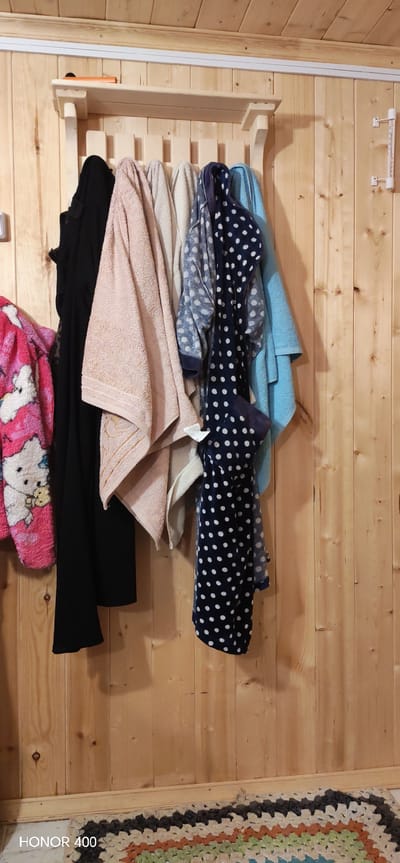 The image depicts a wooden wall with a coat rack holding various items. From left to right, these include a pink robe with a cartoon character, a black garment, a pink towel, a beige towel, a blue robe with white polka dots, and a blue towel. On the right side of the wall is a white thermometer. At the bottom of the image, a multicolored rug is visible. The photograph appears to have been taken indoors. The location is stated as Беркуты, Russia.