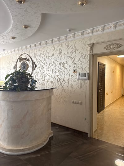 This image depicts an interior scene, likely a hotel lobby or reception area in Dombay, Russia. The camera angle is looking upwards, emphasizing the ornate ceiling and decorative elements. 

The subjects are primarily architectural and decorative objects. A prominent feature is the textured ceiling with a repeating pattern, accented by intricate white and gold molding along the edges. A large, circular clock with an ornate frame hangs on the wall, and below it, a lush green plant in a pot adds a touch of nature to the indoor setting.

Further up, a dark-colored door with a grid pattern is visible, suggesting an entrance or a closed-off area. Next to it, a white intercom or security system is mounted on the wall. There are also light fixtures and wall sockets visible. The flooring transitions from light-colored marble-like tiles to darker, larger tiles, creating a visual separation in the space.

The lighting suggests it is daytime, but there are no windows or indications of the weather outside. The overall impression is one of a well-maintained and elegantly decorated interior space. There is some text visible on a yellow tag hanging from the plant, but it is too blurry to read.