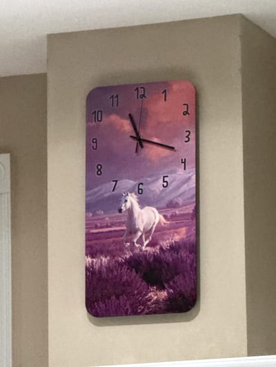 A rectangular clock with rounded corners is mounted on a beige wall. The clock face features a digital rendering of a white horse running through a purple, lavender-like field with mountains and a pink and orange sky in the background. The numbers 1 through 12 are visible on the clock face. The clock has black hour and minute hands, with the hour hand pointing between the 12 and the 1, and the minute hand pointing to the 12. The clock is located in the city of Springboro, United States of America.